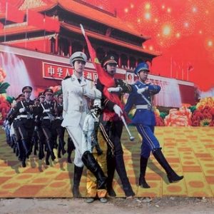NO. 100 – THREE GODDESSES by Liu Bolin, man painted to match the background standing in front of a poster with soldiers