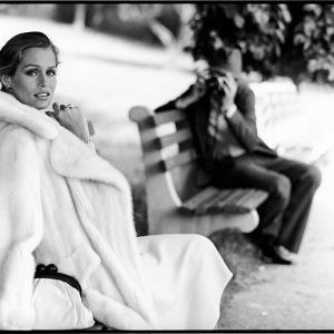 Lauren Hutton for Ultima Revlon. 1975 by Arthur Elgort, model in white fur coat on a parkbench, a man on the bench next to her taking a picture