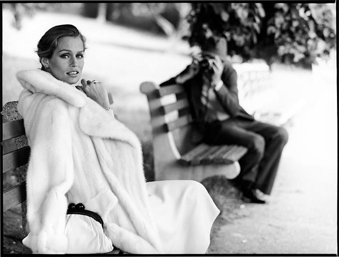 Lauren Hutton for Ultima Revlon. 1975 by Arthur Elgort, model in white fur coat on a parkbench, a man on the bench next to her taking a picture