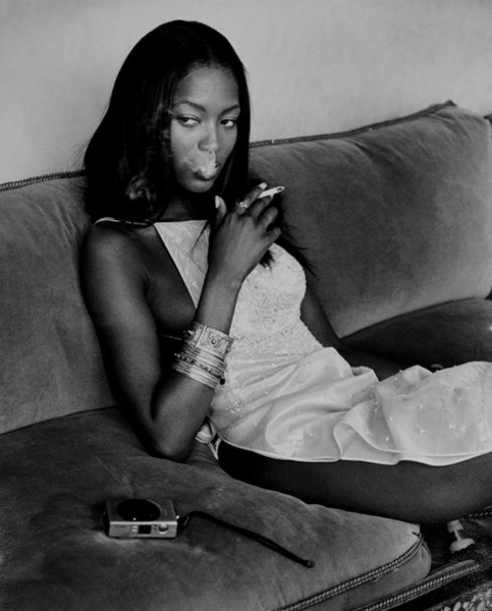 Naomi Campbell Smoking by Albert watson, the model in a white dress sitting on a velvet sofa next to a camera