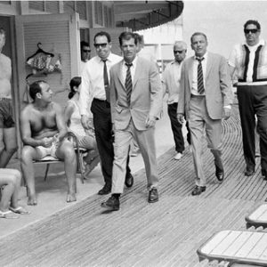 Frank Sinatra Boardwalk, 1968 by Terry O'Neill, group of men in suits walking past people in bathing clothes on sunbenches