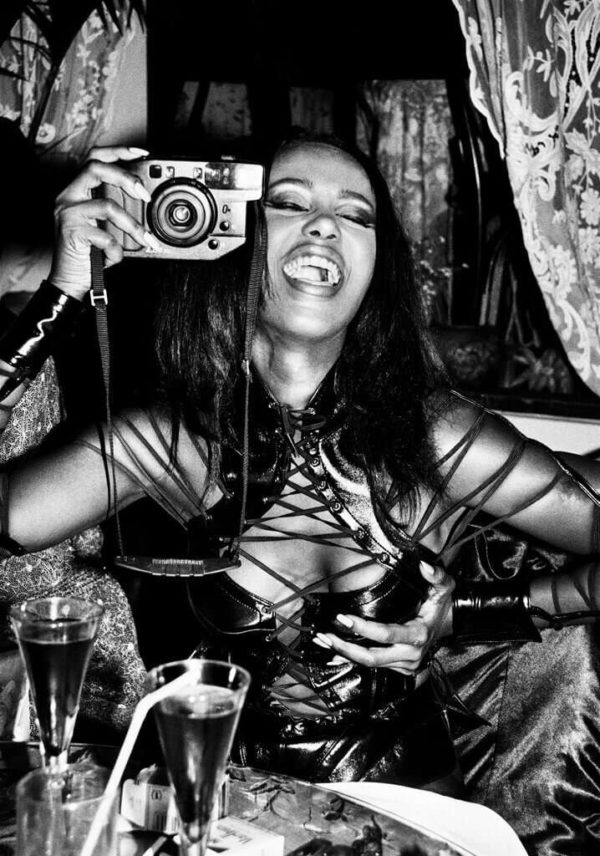 Iman, Paris 1990 by Roxanne Lowit, the model in a black leather bondage top sitting at a table with drinks, holding a camera, laughing