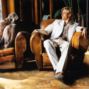Brad Pitt 1997 by Mark Seliger, the actor in a white suit sitting in a leather armchair, a dog sitting in the chair next to him, both looking out the window