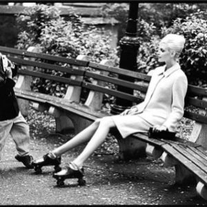 Warren Photographing Nadja. 1995 by Arthur elgort, the model in a white coat and roller skates sitting on a park bench, a boy standing in front of her and taking a picture