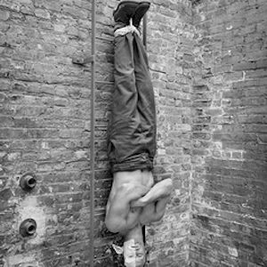 matthew barney II by Mark Seliger, the comedian partially in makeup from his films the cremaster, strung up by his feet in front of a brick wall