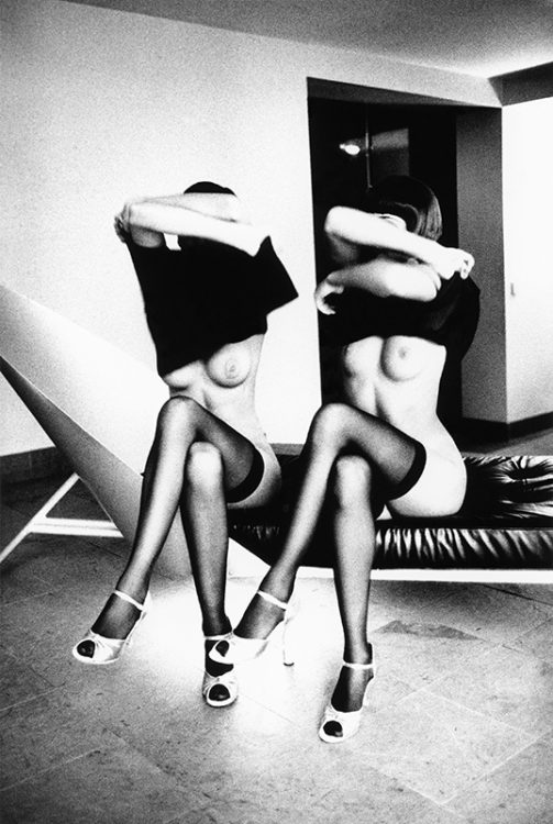 Nudes by Ellen von Unwerth, two modesl in black stockings and white heels sitting on a bench with crossed legs, pulling their shirts up over their heads to expose their breasts