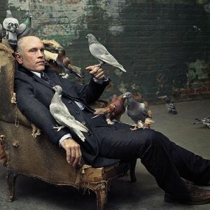 john malkovich with doves by Mark Seliger, the actor in a suit sitting in a broken armchair, surrounded by doves