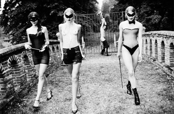 Punishment 2002 by Ellen von Unwerth, three models in dessous and masks, carrying whips, walking away from a man tied to a gardenfence
