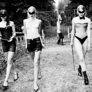 Punishment 2002 by Ellen von Unwerth, three models in dessous and masks, carrying whips, walking away from a man tied to a gardenfence