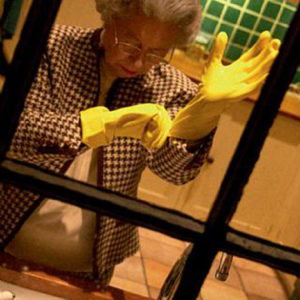 Queen Marigolds by Alison Jackson, Queen lookalike putting on yellow gloves to do the dishes, through a window
