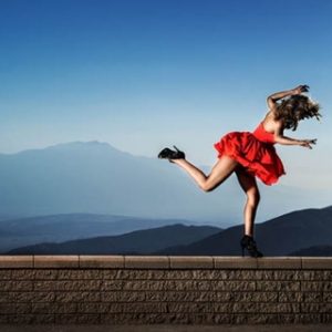 Risky Game 2014 by David Drebin, model in short red dress and black heels stumbleing and balancing on a wall, mountains in the background