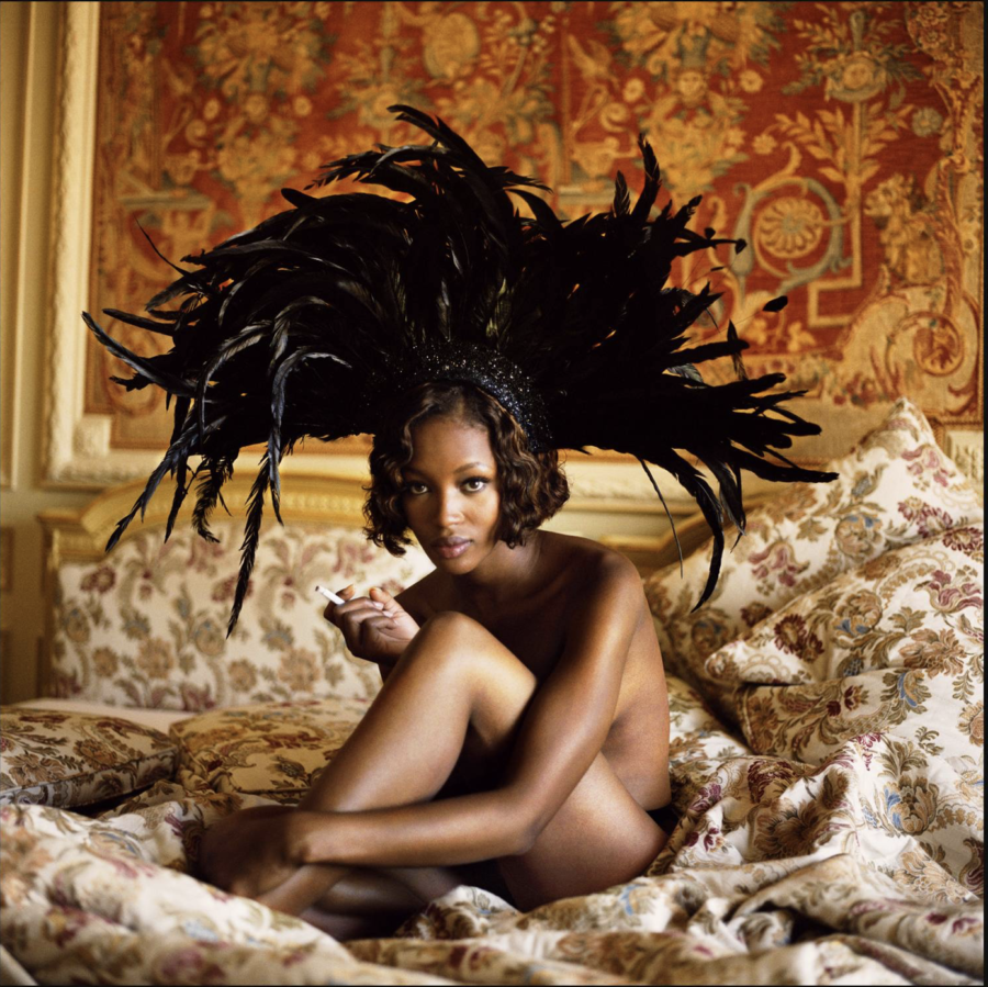 Naomi Campbell, Vogue Italia 1993 by Michel Comte, the nude model sitting in a baroque bed in front of tapestry, wearing a black feather headpiece, smoking