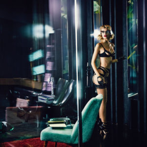 Peep by Ellen von Unwerth, model in black lingerie, heels and gloves standing next to the window in a livingroom