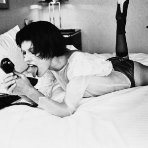 TELEPHONE SEX New York 1997, from the fräulein Series by Ellen von Unwerth, model in white blouse and black tights lying on a hotel bed, licking a telephone