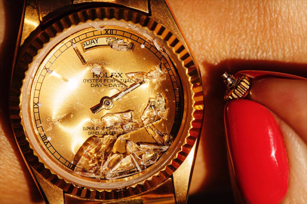 Gold Digger by Tony Kelly, golden Rolex Watch with broken glass and red nails holding a broken off butten next to it