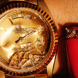 Gold Digger by Tony Kelly, golden Rolex Watch with broken glass and red nails holding a broken off butten next to it