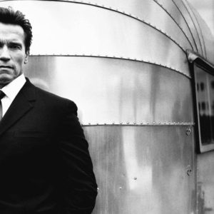 Arnold Schwarzenegger by Nigel Parry, the bodybuilder in a black suit standing in front of a metal caravan