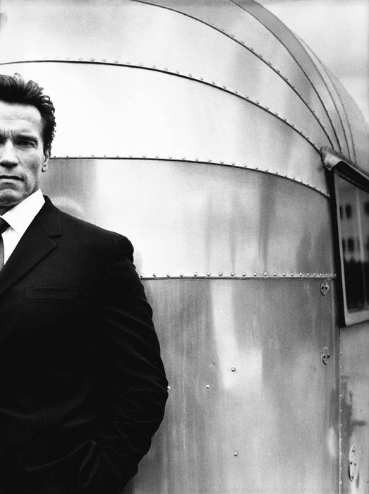 Arnold Schwarzenegger by Nigel Parry, the bodybuilder in a black suit standing in front of a metal caravan