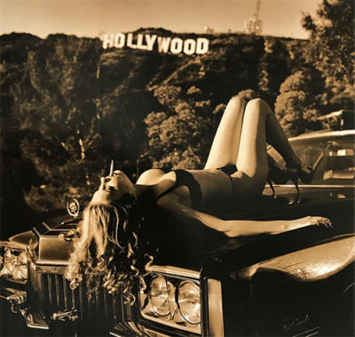 Hollywood Cadillac by Guido Argentini, model in black underwear and heels lying on an engine hood in front of the hollywood sign, smoking