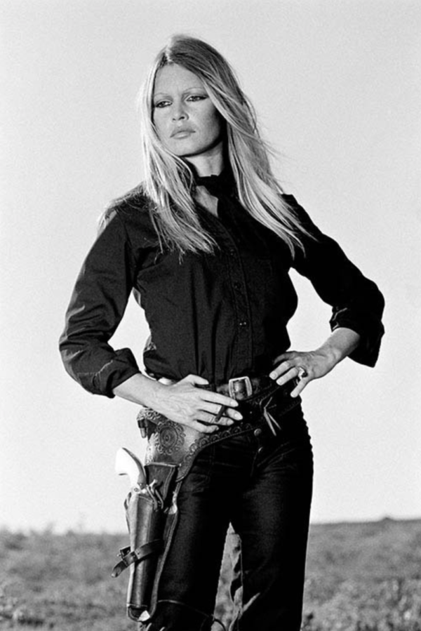 Brigitte Bardot standing by Terry O'Neill, the model in black with a holster and gun, standing with her hands on her hips and a cigarette
