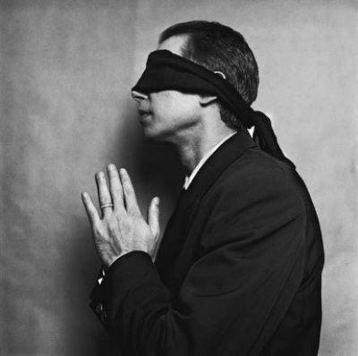 Jeff Koons by Michel Comte, the artist in sideprofile weraing a suit and blindfold with praying gesture