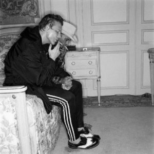 Mickey Rourke by Michel Comte, the actor sitting on a bed in adiadas pants and dress shoes, smoking
