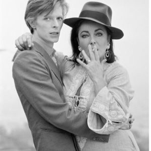 David Bowie & Elizabeth Taylor by Terry O'neill, portrait of Bowie holding Elisabeth who is wearing a fedora and smoking