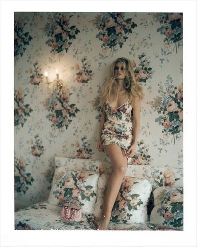Nathalie B. Deauville, blonde model in a short dress, standing on a bed and leaning on a wallpaper wall, all with the dame floral print