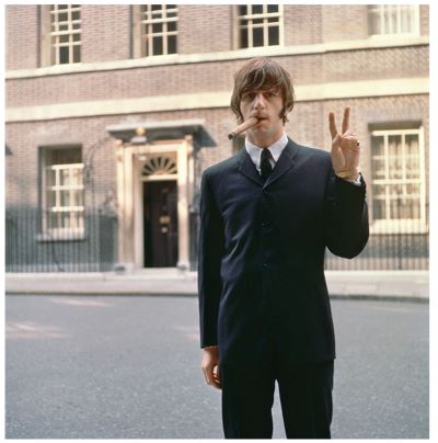 Ringo Starr by Terry O'Neill, the musician in a black suit smoking a giant cigar and showing a piece sign, standing in front of 10 Downing Street