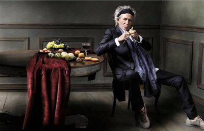 Keith Richards II by Mark Seliger, the singer in a black suit leanig on a table with fruits and red cloth