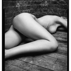 Manon by Mark Seliger, nude model lying on wood floor in front of a brick wall