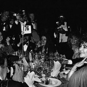 Liz Taylor and Paparazzi, Night of 100 Stars, the actress in a gown sitting at a table surrounded by Photographers