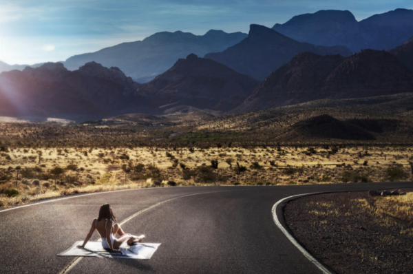 Road Warrior by David Drebin, model in white swimsuit sitting on a white blanket in the middle of the road surrounded by nature