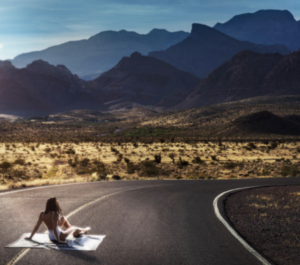 Road Warrior by David Drebin, model in white swimsuit sitting on a white blanket in the middle of the road surrounded by nature