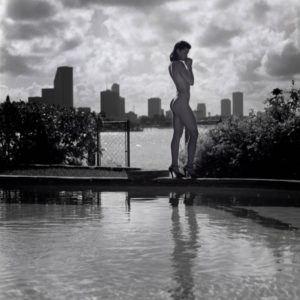 Kim under a cloudy sky by Guido Argentini, nude model in heels standing next to a pool in front of a skyline