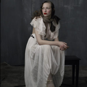 Ala - New York City 2016 by Albert Watson, model in white transparent ruffle gown and red lip, sitting in front of a grey wall