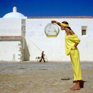Julie Foster - Portugal 1977 by albert watson, model in yellow holding a bird cage, standing in front of a white mediterranen house and blue sky
