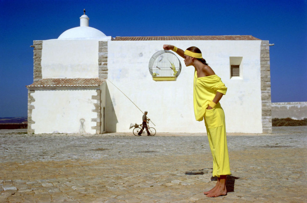 Julie Foster - Portugal 1977 by albert watson, model in yellow holding a bird cage, standing in front of a white mediterranen house and blue sky