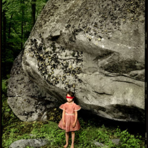 Kylie - Hide n Seek - Sherman - Connecticut 2008 by Albert Watson, a girl in a pink dress and red blindfold standing in a forest next to a huge rock