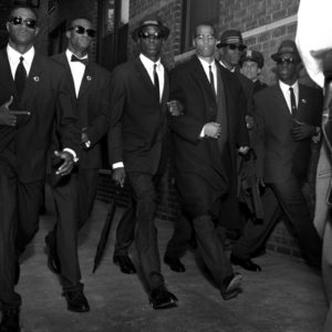 Malcolm X - New York City 1992 by Albert Watson, the civil rights activist in a black suit walking amongst oder suited men in hats and sunglasses, a man wearing a gun in the foreground