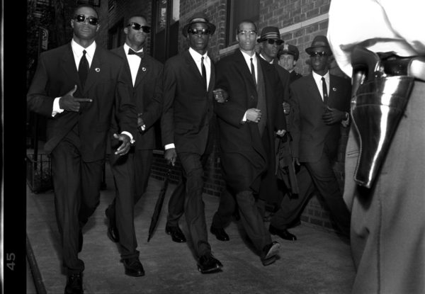 Malcolm X - New York City 1992 by Albert Watson, the civil rights activist in a black suit walking amongst oder suited men in hats and sunglasses, a man wearing a gun in the foreground