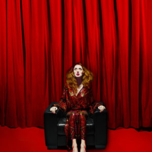 Teddy Quinlivan - Numero Berlin Magazine - NYC 2018 by Albert Watson, the british model in a red sequin jumber, sitting on a black leather armchair in front of red curtains and a red carpet