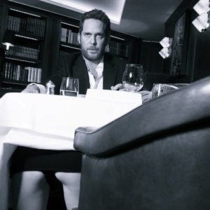 Tom Hollander by Alison Jackson, lookalike of the actor sitting at a table with wine glasses while a woman is kneeling under the table