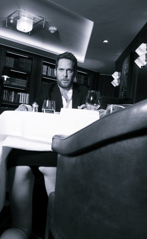Tom Hollander by Alison Jackson, lookalike of the actor sitting at a table with wine glasses while a woman is kneeling under the table