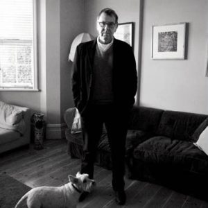 Tom Wilkinson by Alison Jackson, lookalike of the actor in a black suit standing in a living room with a white dog