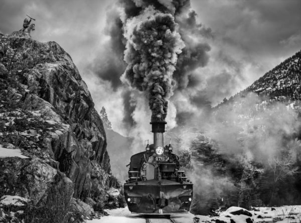 Vantage Point by David Yarrow, steam locomotive with heavy smoke between rocks, a native american with a gun standing on top of a hill looking down