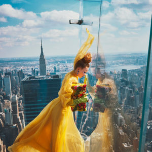 Hadley Robinson IV by Guy Aroch, the model in a yellow gown, green gloves and floral bag, looking through the glass reiling of a skyscrapers rooftop terrace, the city in the background