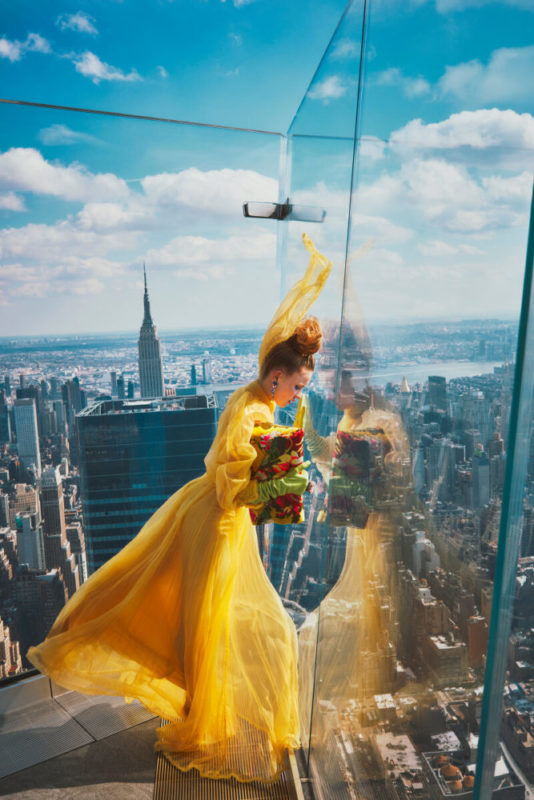 Hadley Robinson IV by Guy Aroch, the actress in a yellow gown, green gloves and floral bag, looking through the glass reiling of a skyscrapers rooftop terrace, the city in the background