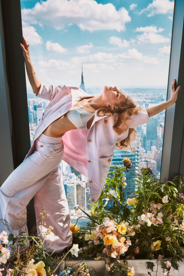 Hadley Robinson V by Guy Aroch, the actress in a light pink suit atanding between flowers in the window of a skyscraper, the city in the background