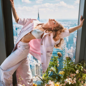 Hadley Robinson V by Guy Aroch, the actress in a light pink suit atanding between flowers in the window of a skyscraper, the city in the background